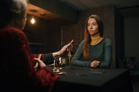 Photo for Young woman asking advice at mature old gypsy taro reader. Female client and fortune teller sitting at table with burning candle - Royalty Free Image