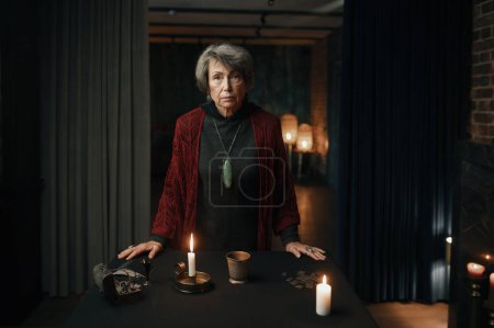 Foto de Portrait of old mature gypsy witch standing at table with psychic tools in mystical atmosphere - Imagen libre de derechos