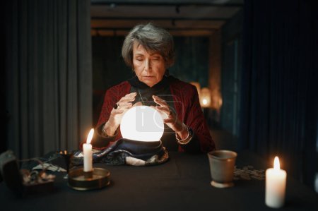 Photo for Portrait of old mature fortune teller looking into illuminated crystal ball to predict future and divination - Royalty Free Image