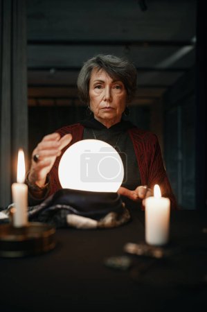 Photo for Confident witch fortune teller doing predictions with illuminated crystal ball reading future during esoteric ritual - Royalty Free Image