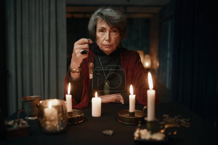 Téléchargez les photos : Portrait of serious confident fortune teller holding crystal pendant over candle light for divination seance - en image libre de droit