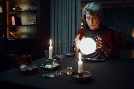 Photo for Confident witch fortune teller doing predictions with illuminated crystal ball reading future during esoteric ritual and divination seance - Royalty Free Image