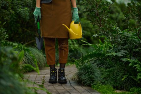 Photo for Cropped shot of female gardener dressed in apron holding garden tool in hands wearing rubber gloves view on leg in boots over greenhouse background - Royalty Free Image