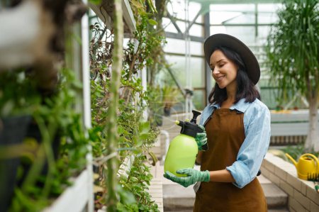 Foto de Young woman gardener caring plants treating flowers with chemicals from pulverizer sprayer. Greenhouse work - Imagen libre de derechos