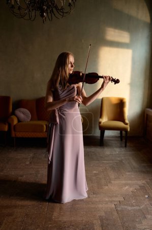Foto de Beautiful woman wearing elegant dress playing violin standing along in home room - Imagen libre de derechos