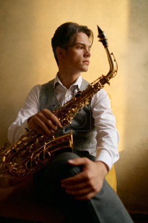 Photo for Young confident man rest in chair with saxophone under sunlight spreading from window. Jazz musician cradling sax - Royalty Free Image