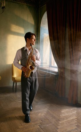 Téléchargez les photos : Young man practicing saxophone at home during weekend leisure time. Happy smiling saxophonist full-length portrait - en image libre de droit