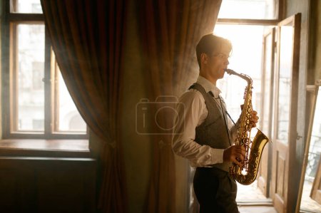 Téléchargez les photos : Young elegant man playing gold alto saxophone in misty room. Emotional musician with sax - en image libre de droit