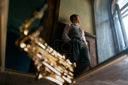 Téléchargez les photos : Pensive young saxophonist looking through window view from bottom, selective focus mirror reflection - en image libre de droit