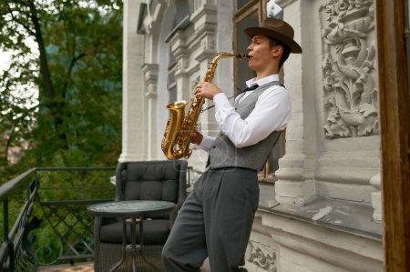 Téléchargez les photos : Young male musician practicing saxophone in balcony at home. Inspired saxophonist playing music - en image libre de droit