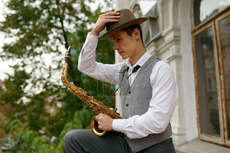 Téléchargez les photos : Portrait of young musician wearing heat and stylish outfit holding saxophone musical instrument. Handsome jazzman looking at camera - en image libre de droit