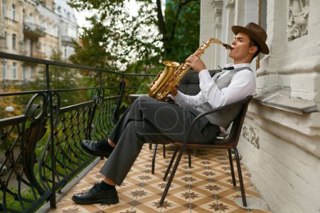 Téléchargez les photos : Handsome jazzman enjoys playing saxophone relaxing on comfortable home terrace - en image libre de droit