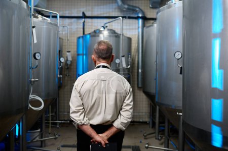 Téléchargez les photos : Back view of brewer man in apron standing among distillery vats at beer manufacturing factory - en image libre de droit