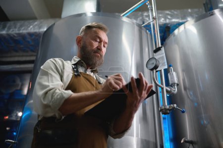 Téléchargez les photos : Handsome bearded man brewer inside modern beer factory around steel tanks. Mature male worker making notes about technological process - en image libre de droit