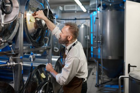 Téléchargez les photos : Concentrated male brewer making notes on the tanks during brewing process. Craft beer manufactory concept - en image libre de droit