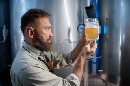 Foto de Brewery worker looking at freshly made beer in glass mug. Brewing factory technological process concept - Imagen libre de derechos