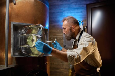 Téléchargez les photos : Homme brasseur examinant réservoir de bière processus d'inspection de brassage. Couvercle d'ouverture de la bouilloire métallique et regard vers le bas, confiant et barbu - en image libre de droit