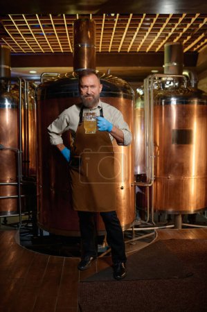 Téléchargez les photos : Senior man brewery technologist holding fresh craft beer at microbrewery plant. Inspector working at alcohol manufacturing factory checking ale or lager - en image libre de droit