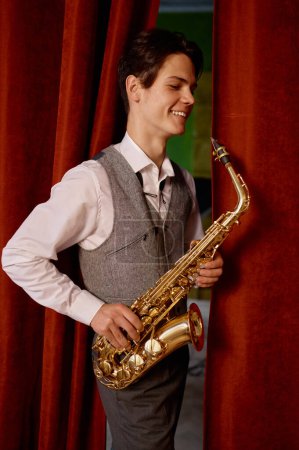 Téléchargez les photos : Smiling jazz saxophone player training before performance. Satisfied saxophonist standing over red stage curtains - en image libre de droit