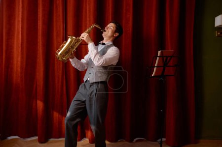 Photo for Young attractive musician plays tenor saxophone on stage with red drape curtains. Jazz festival - Royalty Free Image