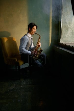 Photo for Jazz musician playing saxophone in front of window sitting in chair in retro room - Royalty Free Image