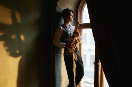 Foto de Young man with saxophone standing on windowsill of old room in daylight. - Imagen libre de derechos