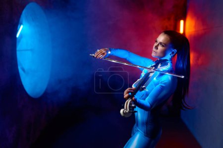 Téléchargez les photos : Beautiful violinist wearing tight tracksuit playing classic music in neon light. Futuristic, sci-fi loft design - en image libre de droit