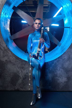 Téléchargez les photos : Young woman playing magic melody on electronic violin in neon glowing light standing against industrial air blower turbine - en image libre de droit