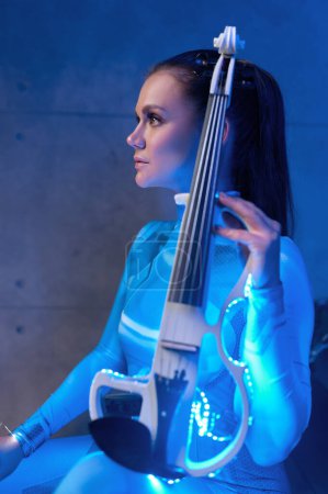Photo for Side view of young woman violinist with electric fiddle in neon glowing light. Futuristic concert - Royalty Free Image