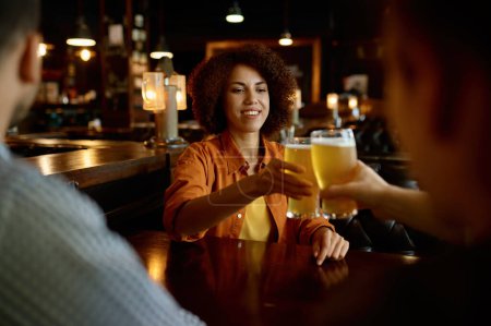 Photo for Friends drinking beer cheering glasses sitting at table in sport bar. Focus on happy smiling woman. Friendship, rest and recreation concept - Royalty Free Image