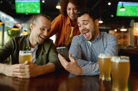 Foto de Cheerful young people rest in sport bar. Laughing friends sitting at table and watching funny video on smartphone while drinking draft beer - Imagen libre de derechos
