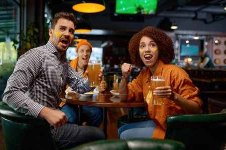 Téléchargez les photos : Cheering friends of soccer fans in pub. Multiracial diverse group of young people with funny makeup in faces watching game on screen in sports bar cheering for favorite team during football festival - en image libre de droit