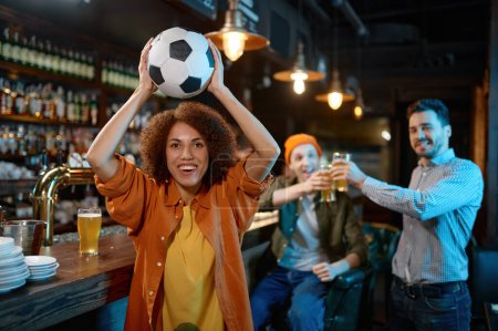 Téléchargez les photos : Happy friends rest at sports bar and drinking beer. Focus on overjoyed young woman holding soccer ball over head ready to throw into camera - en image libre de droit