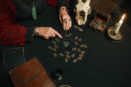 Photo for Old mature witch giving prediction using spiritual attributes. Closeup top view on sorceress hand moving crystal pendulum and metal runes - Royalty Free Image