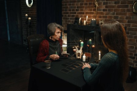 Téléchargez les photos : Female enchantress witch spreading tarot cards at table front of young woman client to predict the future. Divination concept - en image libre de droit