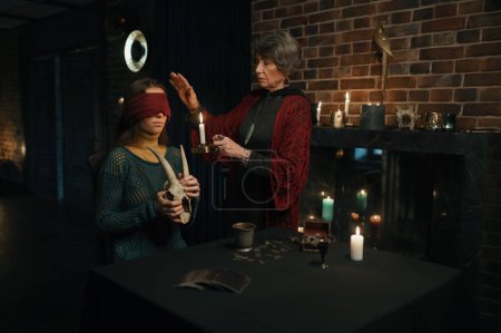 Photo for Old mature witch sorceress holding magic ritual for young female client. Wise woman blindfolding girl standing in atmospheric witchcraft office - Royalty Free Image