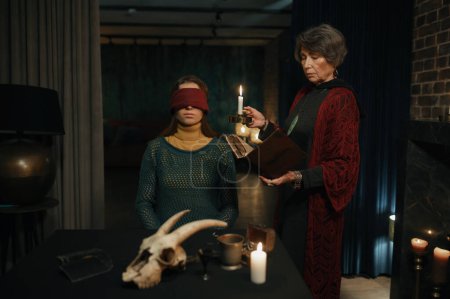Téléchargez les photos : Old mature gypsy fortune teller reading witchcraft spell from book for blindfolding woman. Sorceress holding magic ritual spiritual session - en image libre de droit