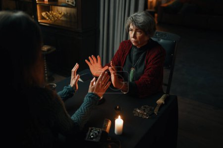 Photo for Fortune teller sorceress and woman sitting at table desk doing spiritual ritual over candle flame - Royalty Free Image