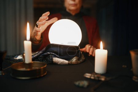 Photo for Confident witch fortune teller doing predictions with illuminated crystal ball reading future during esoteric ritual and divination seance - Royalty Free Image