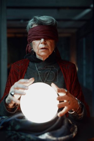 Photo for Blindfolded witch fortune teller doing predictions with illuminated crystal ball reading future during esoteric ritual and divination seance - Royalty Free Image