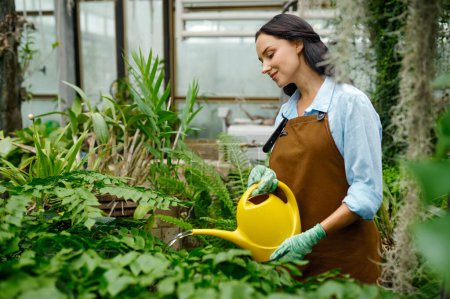 Foto de Young woman florist caring flower plants watering green shoots from plastic can at greenhouse. Farming or gardening concept - Imagen libre de derechos
