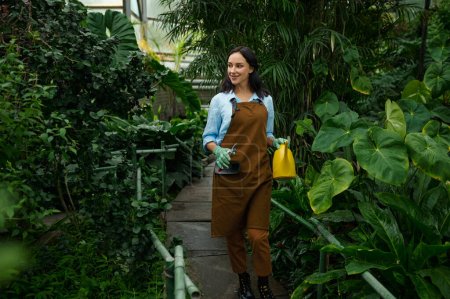 Téléchargez les photos : Young woman gardener working in botanic garden. Millennial girl florist walking through lush green plants in greenhouse - en image libre de droit