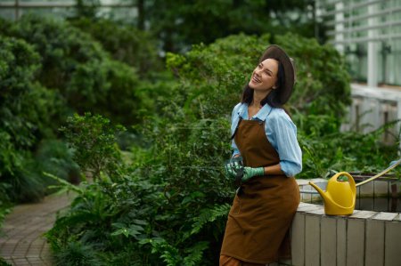 Téléchargez les photos : Portrait of positive smiling woman gardener wearing apron carrying flowerpots in hand selective focus with view through lush greenhouse foliage - en image libre de droit