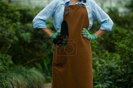 Foto de Closeup female gardener in overalls with garden small shovel and rake tool in pocket. Gardening and farming hobby concept - Imagen libre de derechos