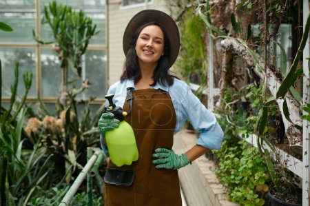 Photo for Portrait of positive smiling young woman gardener with pulverizer sprayer in hand posing for camera over greenhouse garden background - Royalty Free Image