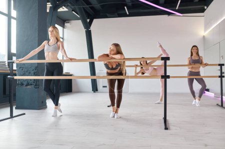 Foto de Young elegant women dancers stretching legs, practicing ballet movement at barre. Training at dance studio gym - Imagen libre de derechos