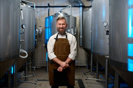 Foto de Retrato del hombre cervecero sonriente en delantal parado entre cubas de destilería en la fábrica de fabricación de cerveza - Imagen libre de derechos