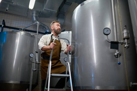Foto de Hombre maduro cervecero escalando escalera contra recipiente de metal en fábrica de cerveza. Concepto de pequeña cervecería - Imagen libre de derechos