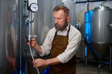 Photo for Adult bearded man in workshop of modern industrial brewery. Male brewer checking brewing mechanism of stainless steel vessel with pressure gauge valve - Royalty Free Image