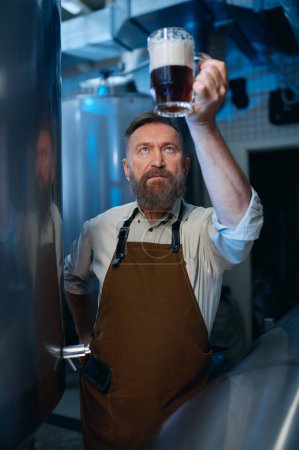 Téléchargez les photos : Bearded senior brewer checking beer quality during brewing technology process. Mature brew master looking at foam level in glass - en image libre de droit